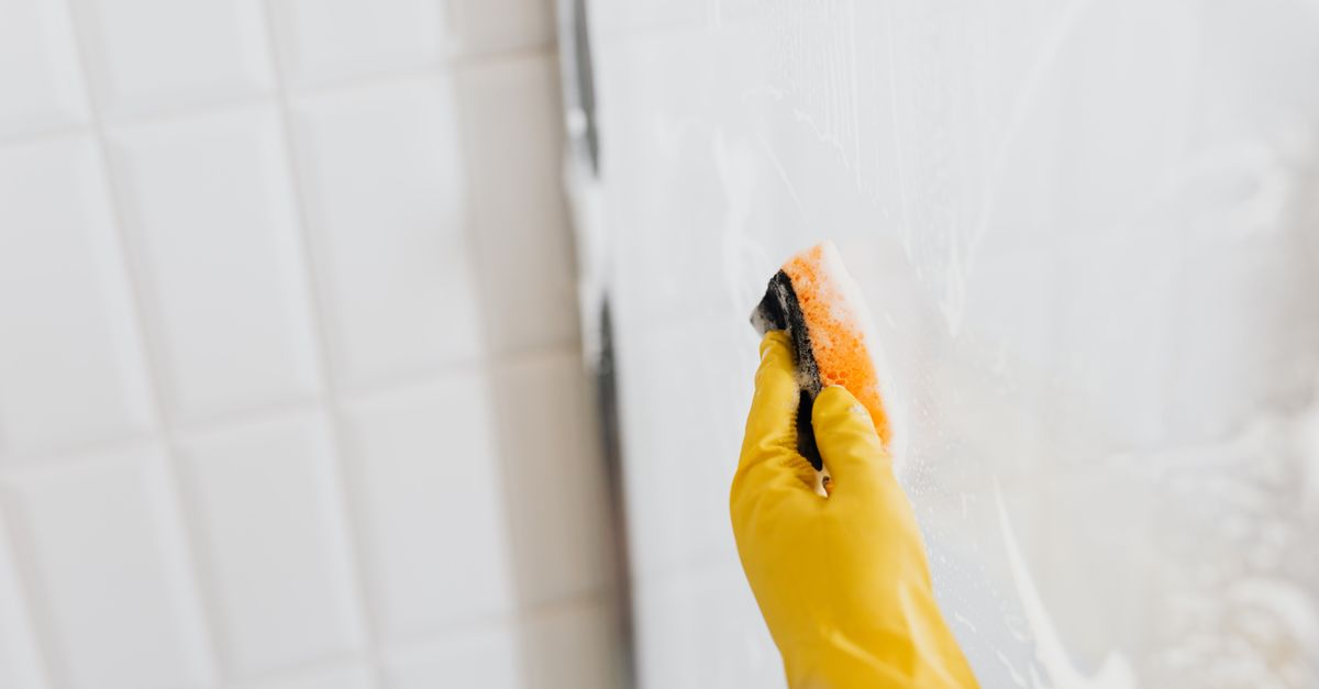 Mão com luva amarela passando esponja com espuma no vidro do box, ao lado de parede de azulejos brancos com rejunte visível.