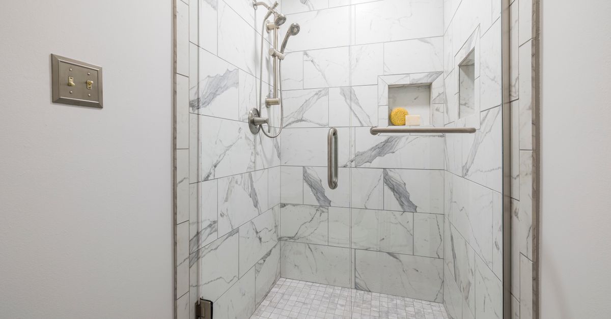 Box de vidro com paredes revestidas de azulejos brancos com veios cinza e rejunte claro, mostrando ducha e nicho para produtos de banho.