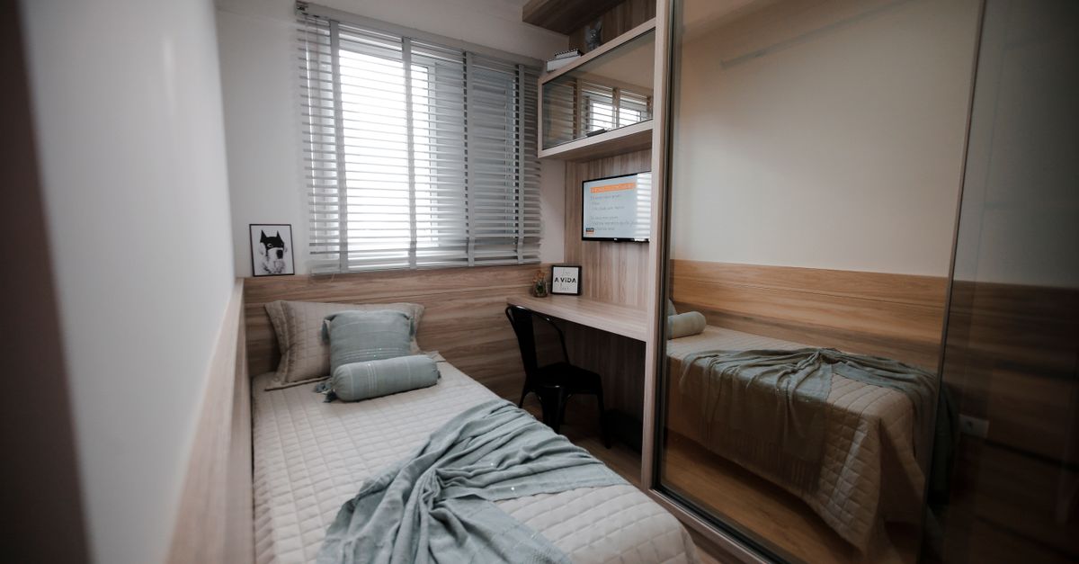 Quarto pequeno e organizado com cama de casal, armário com porta de espelho, escrivaninha embutida e janela com persiana deixando entrar luz natural.
