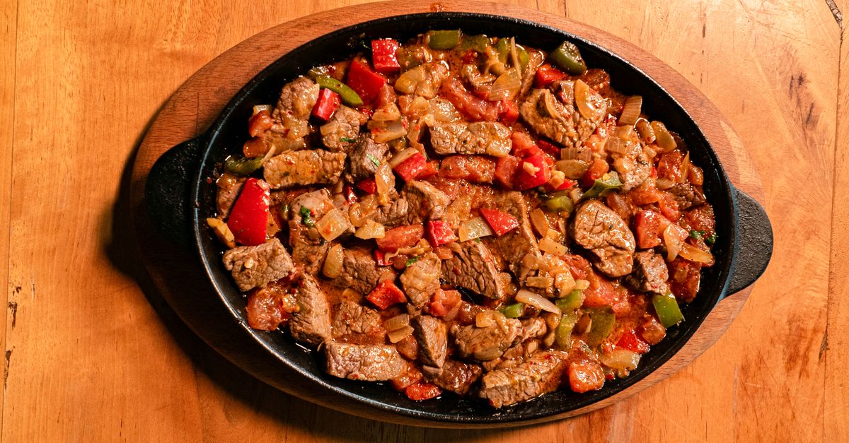 Carne em cubos cozida com molho de tomate, cebola e pimentões servida em uma travessa de ferro fundido sobre suporte de madeira.