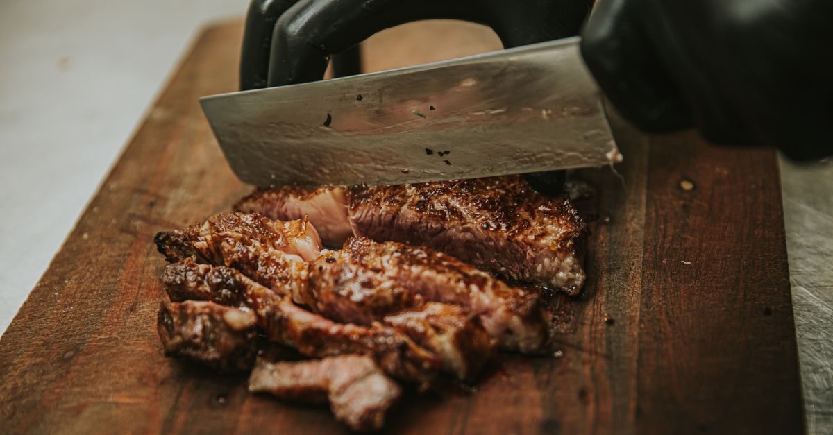 Mão com luva preta segurando uma faca cortando uma peça de carne grelhada em fatias sobre uma tábua de madeira, mostrando as fibras da carne.