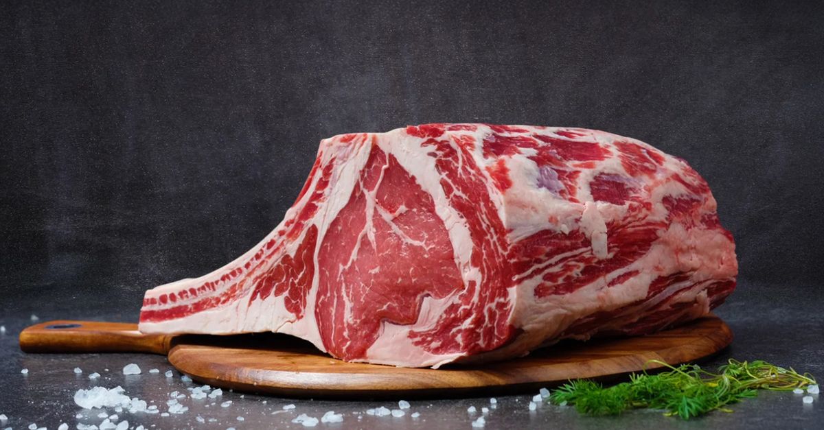 Peça grande de carne bovina crua com marmoreio, temperada com sal grosso e ervas, sobre uma tábua de madeira.
