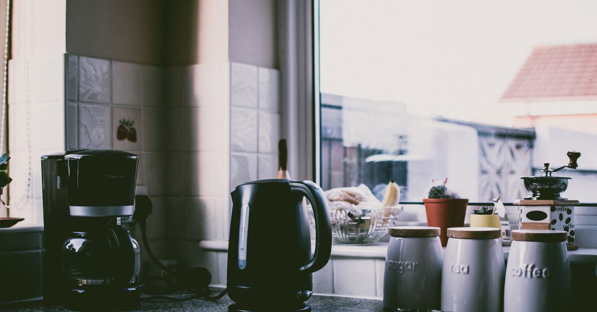 Chaleira elétrica preta sobre a bancada da cozinha, ao lado de uma cafeteira e potes de mantimentos próximos à janela.