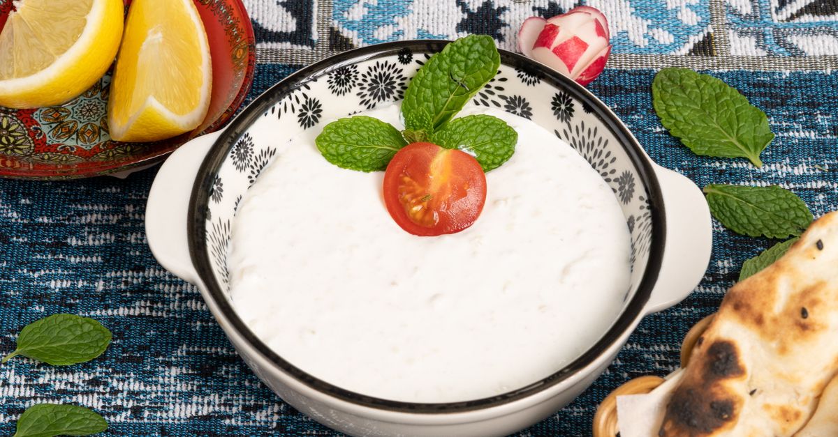 Tigela de creme branco com folhas de hortelã e um tomate cereja no centro, acompanhada de fatias de limão e pão sírio ao lado, sobre toalha azul estampada.