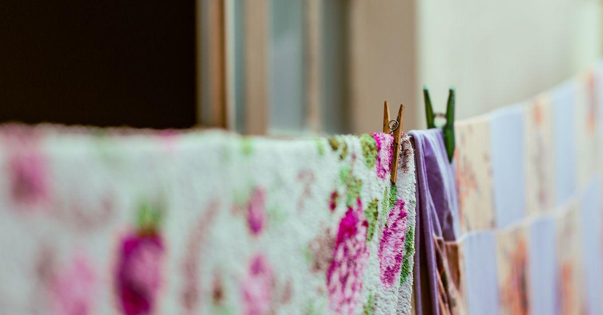 Roupas de tecido floral e lilás penduradas em um varal próximo a uma janela, presas com um pregador de madeira
