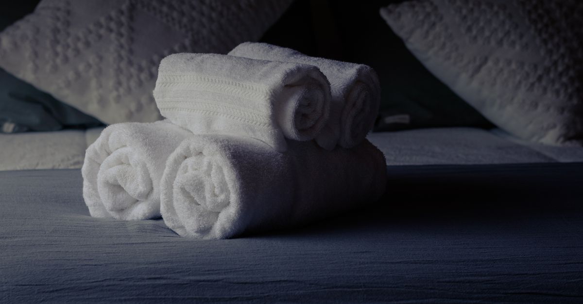 Toalhas brancas macias enroladas e empilhadas sobre a cama, com travesseiros ao fundo.