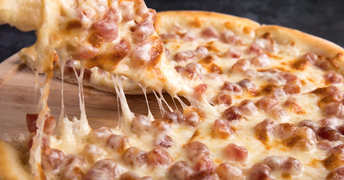 Fatia de pizza sendo levantada com queijo derretido esticando em fios sobre a pizza inteira com pedaços de bacon.