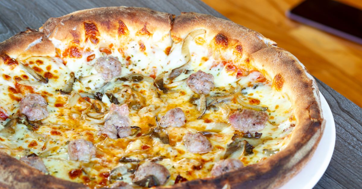 Pizza inteira com borda dourada e crocante, coberta com carne moída, queijo derretido e ervas, servida em prato branco.