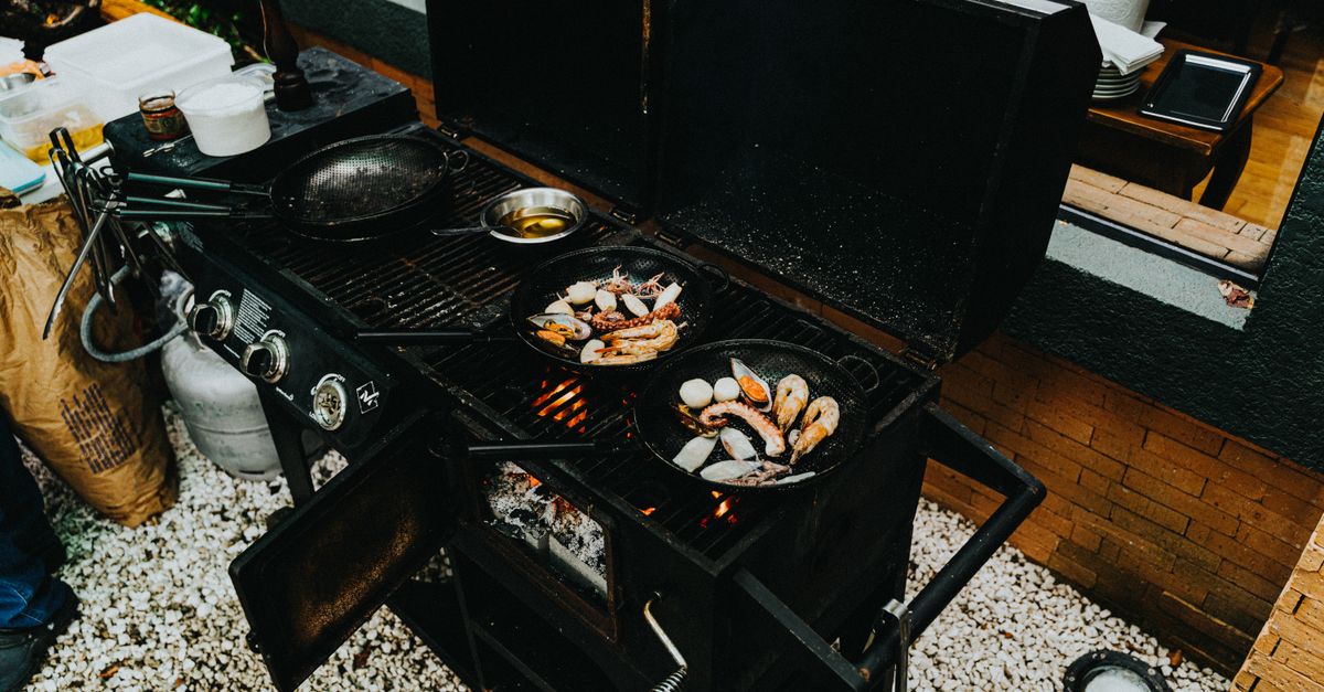 Duas frigideiras de ferro fundido com frutos do mar grelhando sobre um fogão a gás de churrasqueira ao ar livre, com chamas visíveis e outra frigideira vazia ao lado.