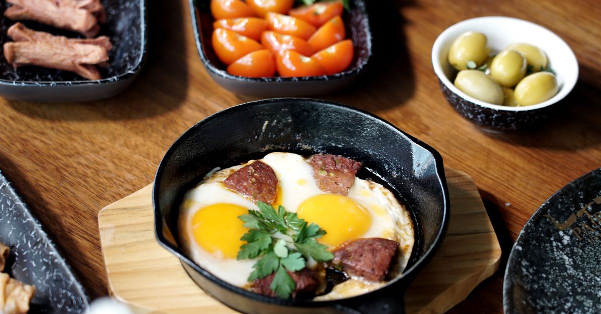 Mini frigideira de ferro fundido com ovos fritos e linguiça, servida sobre tábua de madeira ao lado de acompanhamentos.