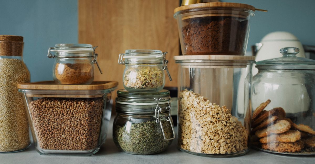 Potes de vidro herméticos com tampas de bambu contendo grãos, cereais e outros alimentos organizados sobre uma bancada de cozinha.