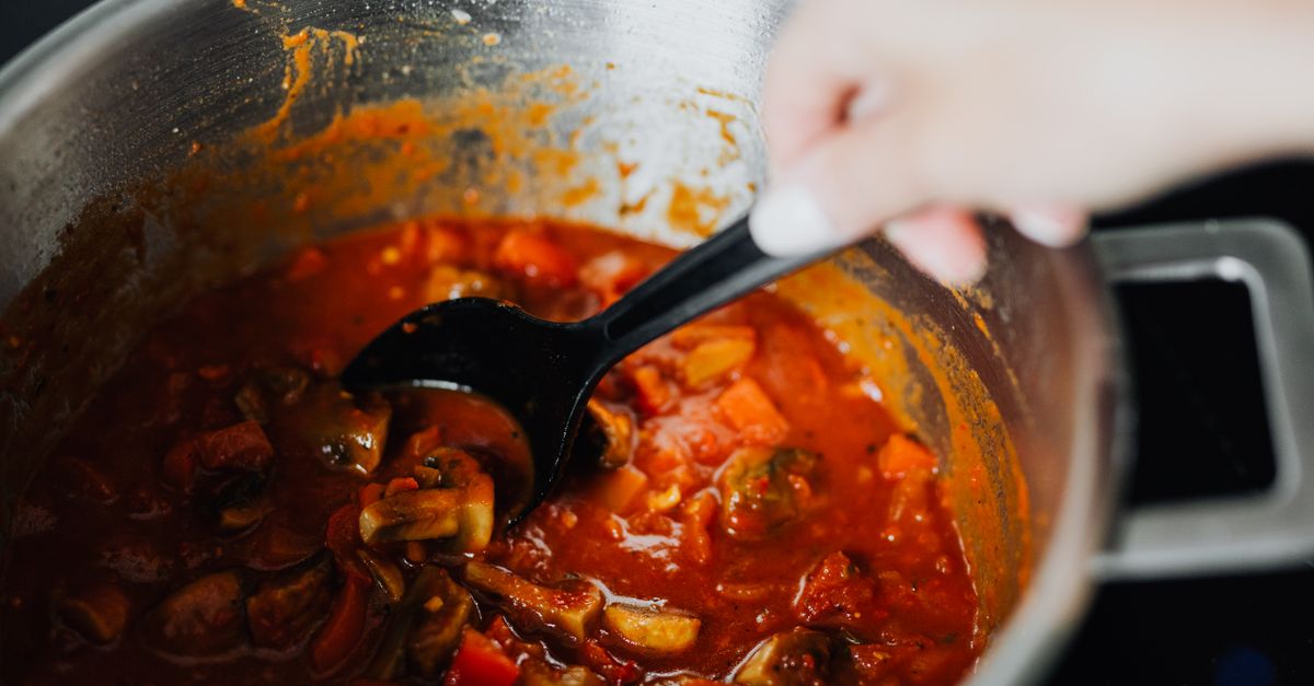 Uma panela com molho de tomate e legumes fervendo no fogão, sendo mexida com uma colher preta.