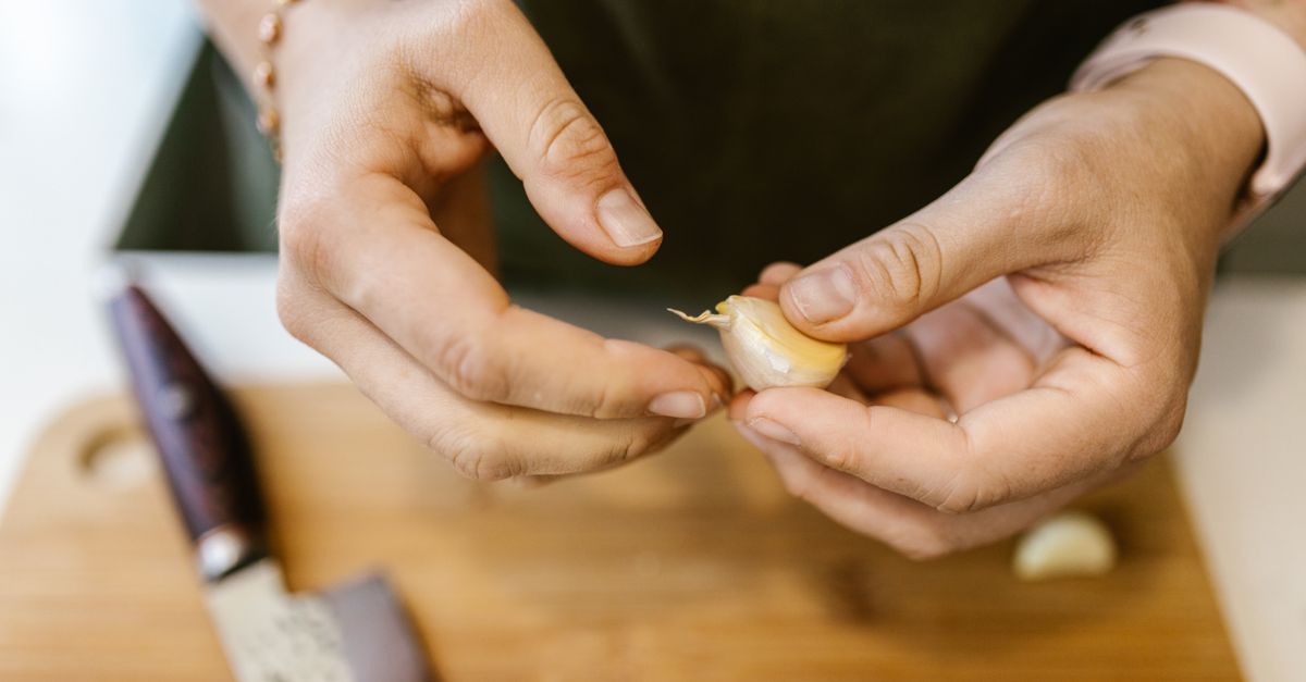 Mãos descascando um dente de alho sobre uma tábua de madeira, com uma faca ao lado.