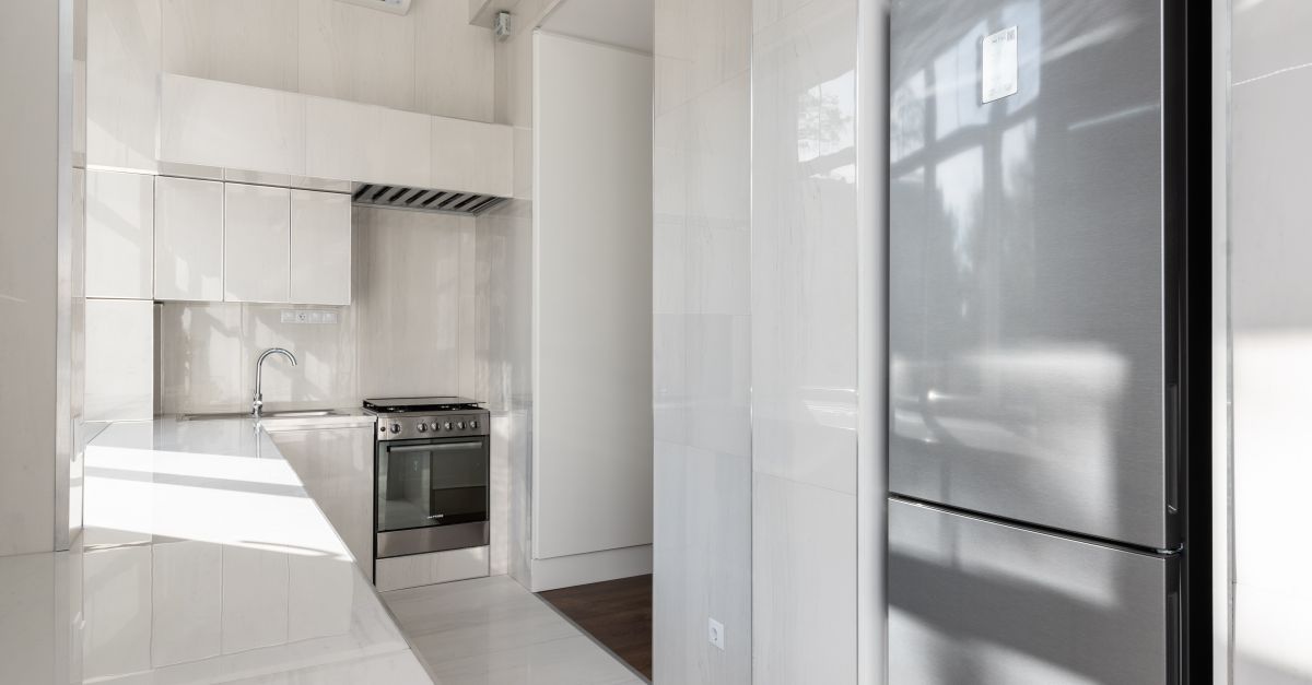 Geladeira de aço inox fechada em cozinha branca e moderna, com fogão e coifa ao fundo.