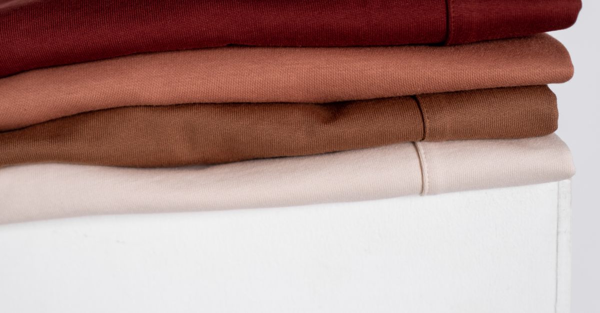 Pilha de lençóis dobrados em tons de vermelho, terracota, marrom e branco, organizados sobre uma superfície branca.