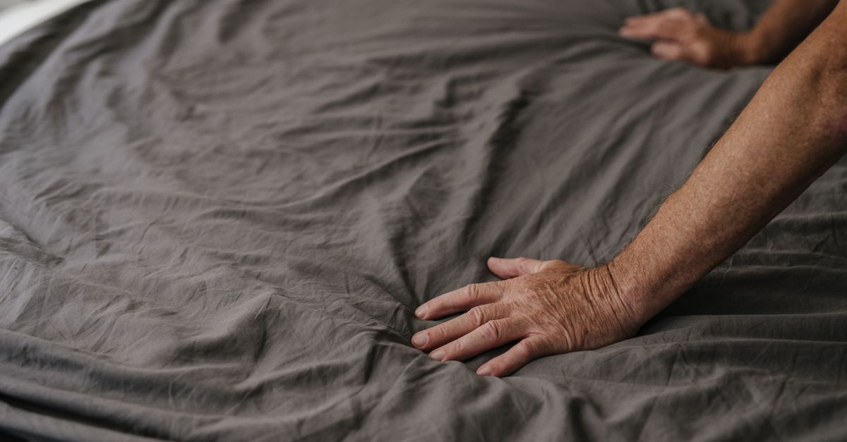 Mão de uma pessoa alisando um lençol cinza amassado sobre a cama.