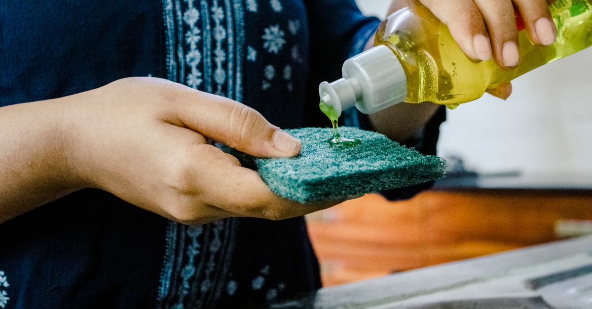Uma pessoa despeja detergente líquido sobre uma esponja verde para esfregar, segurando o frasco na mão.