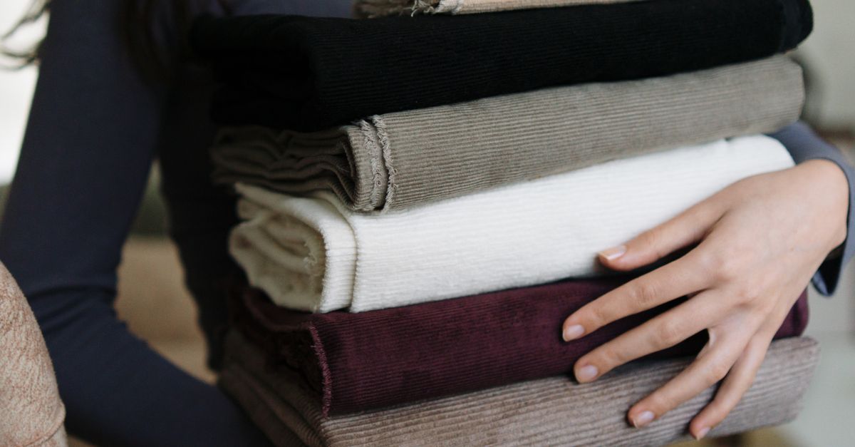 Mulher segurando uma pilha organizada de roupas dobradas de diferentes tecidos, incluindo malha, veludo cotelê e algodão.