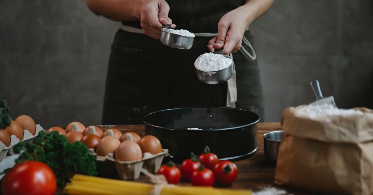 Uma pessoa segura duas xícaras medidoras cheias de farinha sobre uma mesa com ovos, tomates, macarrão e uma forma de bolo.