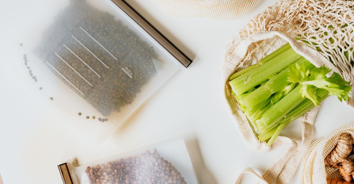 Sacos plásticos com grãos e vagens de vegetais organizados sobre uma superfície branca, ao lado de talos de aipo em uma sacola de tecido.