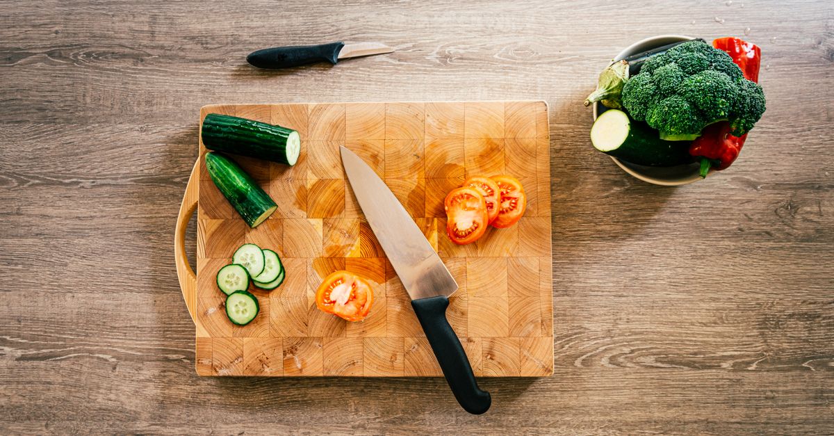 Tábua de madeira com pepino e tomate fatiados, faca ao centro e uma tigela com brócolis, abobrinha e pimentão ao lado.