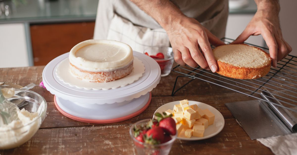Cubos de manteiga em um pratinho branco sobre a mesa de madeira, enquanto uma pessoa monta um bolo ao fundo.