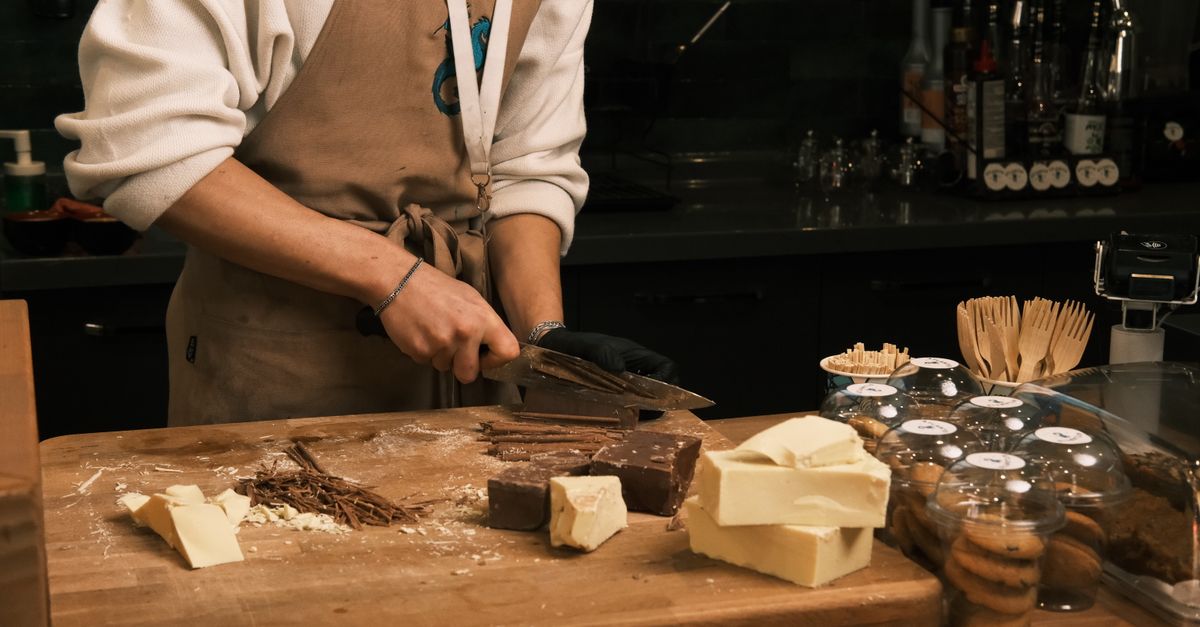 Uma pessoa de avental corta blocos de manteiga e chocolate sobre uma tábua de madeira na cozinha.