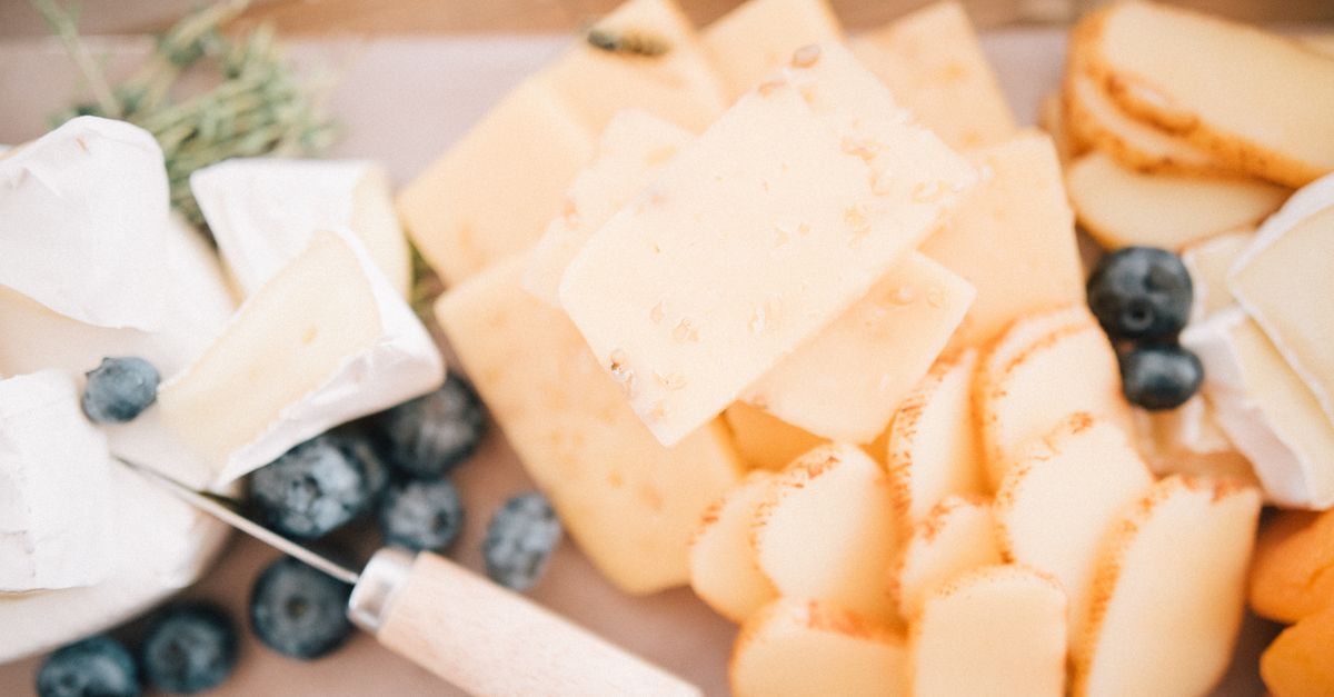 Tábua com cubos e fatias de queijos variados, incluindo queijo cremoso branco, queijo amarelo com furos e queijo curado, acompanhados de mirtilos e um raminho de alecrim.