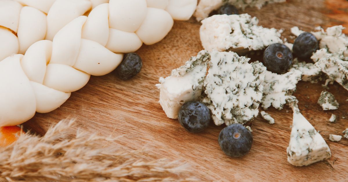 Queijos artesanais, incluindo um trançado e um azul esfarelado, dispostos sobre uma tábua de madeira com mirtilos ao redor.