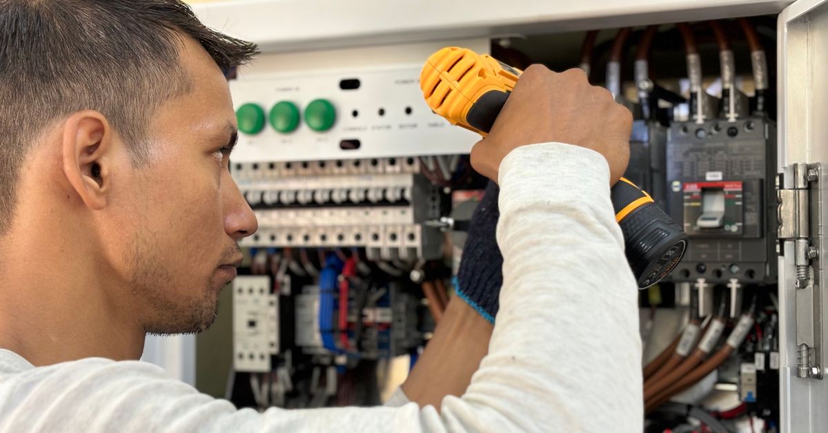 Um eletricista utiliza uma furadeira para trabalhar em um quadro elétrico aberto com disjuntores e fiação organizada.