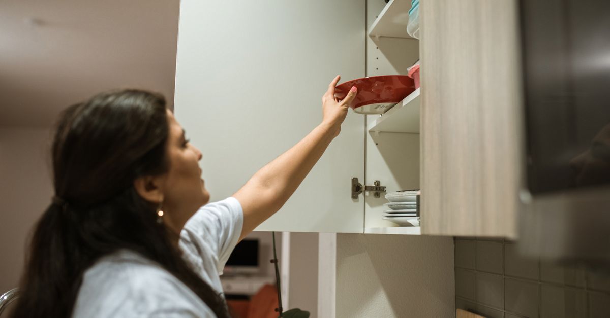 Uma mulher abre um armário de cozinha e organiza pratos e uma tigela vermelha nas prateleiras.