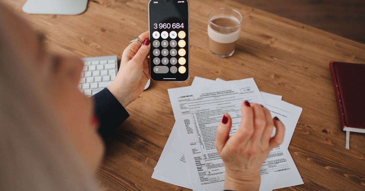 Mulher sentada à mesa de madeira segurando o celular com calculadora aberta enquanto organiza documentos impressos ao lado de um copo de café e um caderno.