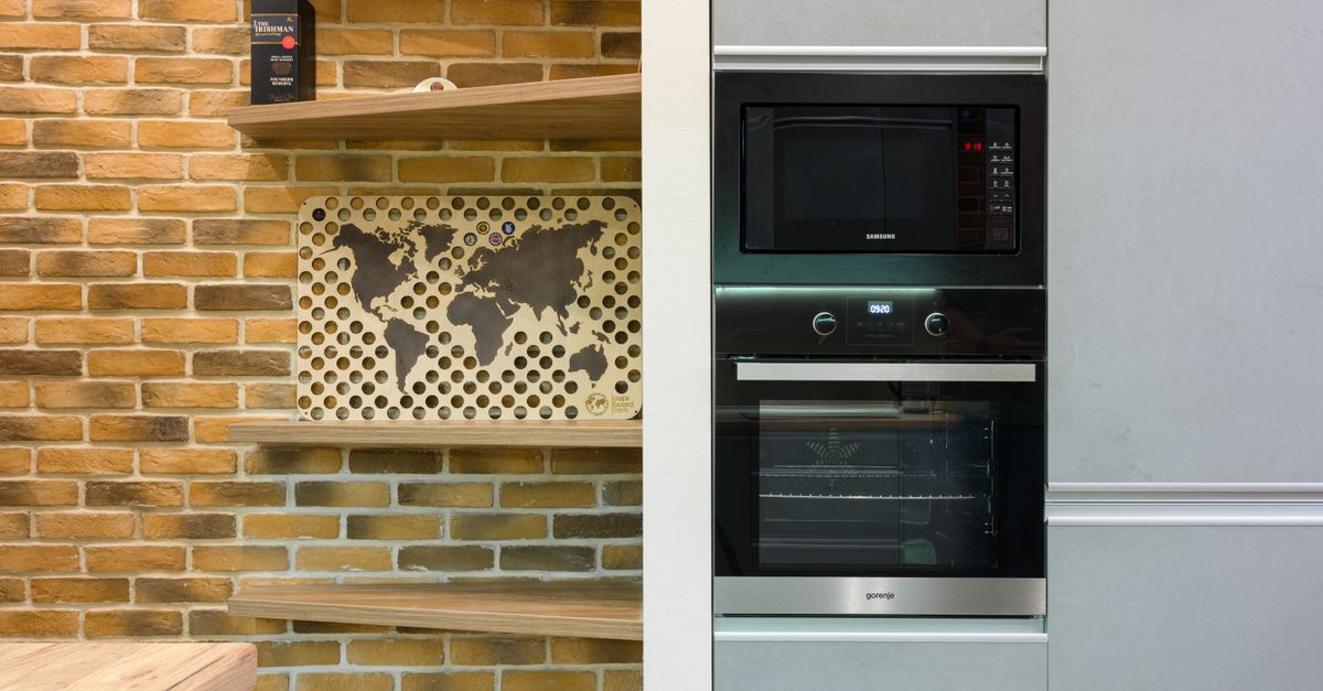 Micro-ondas embutido de cor preta instalado acima de um forno em uma cozinha moderna com parede de tijolinhos ao lado.