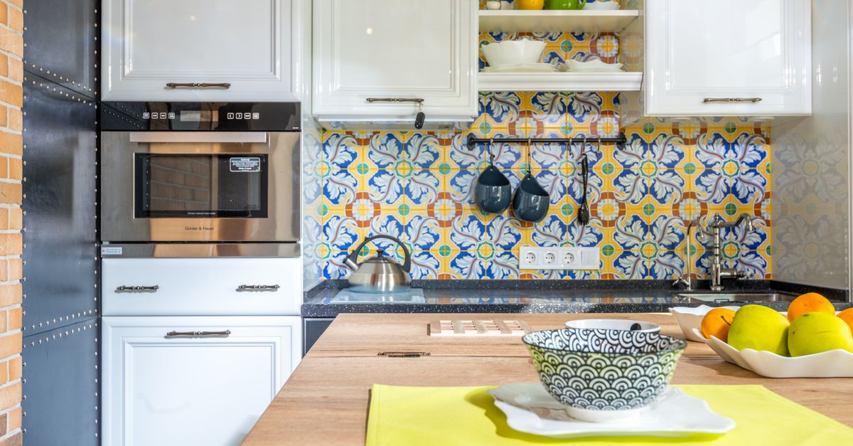 Cozinha com micro-ondas embutido sob armários brancos e azulejos coloridos, com uma tigela e frutas sobre a bancada.
