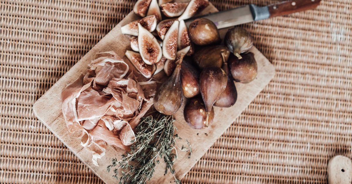 Faca de cozinha ao lado de figos cortados, presunto fatiado e um raminho de ervas sobre uma tábua de madeira.