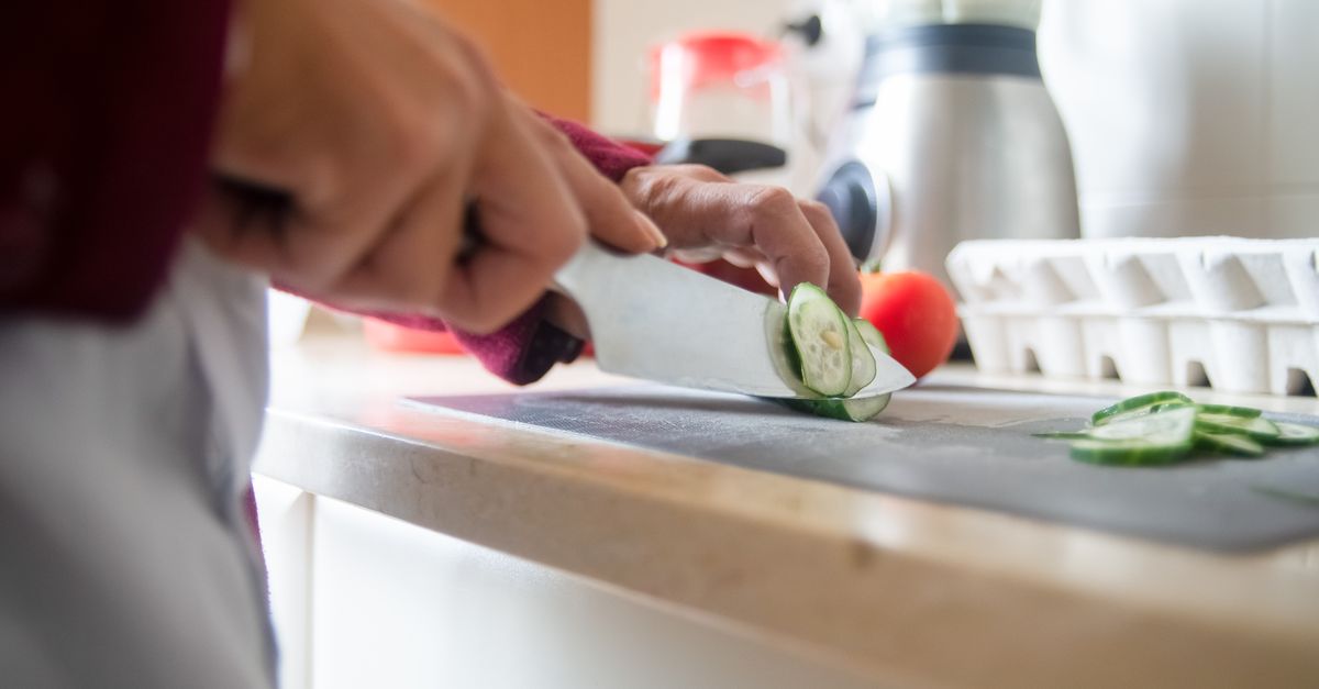 Uma pessoa corta pepino em fatias com uma faca de cozinha sobre uma tábua de corte, com tomate e uma caixa de ovos ao fundo.