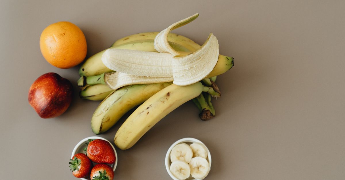 Bananas descascadas e fatiadas em uma tigela branca, ao lado de um cacho de bananas e outras frutas sobre a mesa.