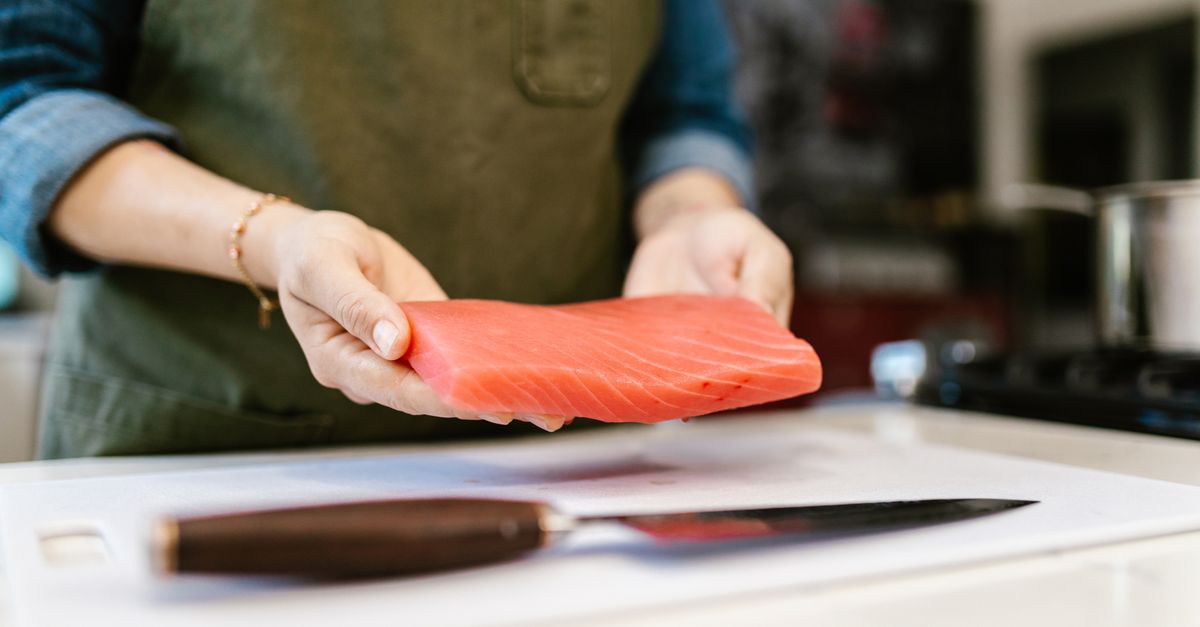 Mãos segurando um filé de peixe cru sobre uma tábua de corte branca, ao lado de uma faca, em uma cozinha.