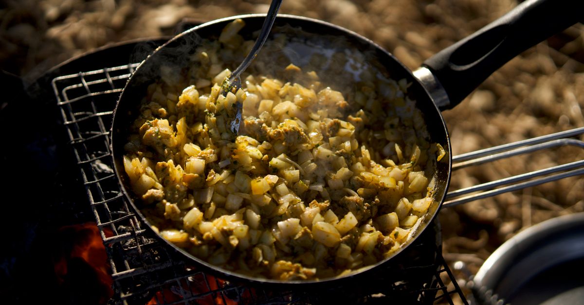 Cebola e legumes picados sendo refogados em uma frigideira sobre fogo aberto, com uma colher mexendo o preparo.