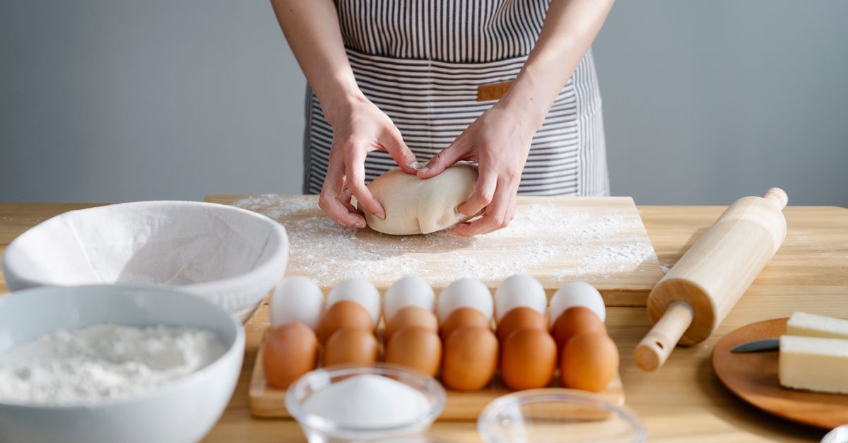 Uma pessoa de avental listrado sova uma bola de massa de pão sobre uma tábua enfarinhada, cercada por ovos, farinha e um rolo de massa.