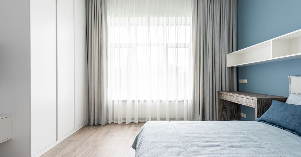 Quarto organizado com parede azul, cortina de voil branca deixando entrar luz natural pela janela, cama com roupa de cama azul e armário branco ao lado.
