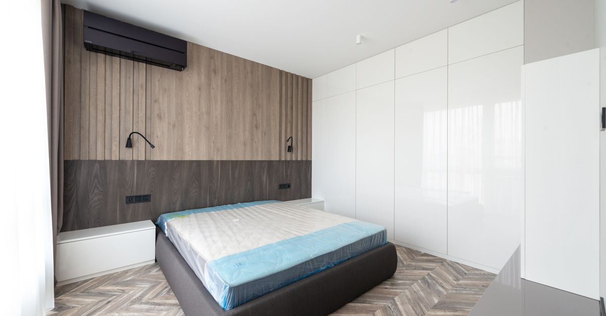 Ar-condicionado split instalado na parede de madeira de um quarto moderno, acima de uma cama com colchão exposto.