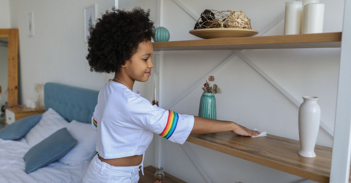 Uma menina limpa uma prateleira de madeira no quarto com um pano, ao lado de vasos e objetos decorativos, com a cama ao fundo.